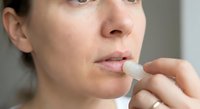 Young woman applying lip balm stick to moisturize dry chapped lips in natural light. Skincare routine and lip care concept for health and beauty.