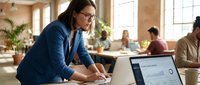 Professional businesswoman with glasses working on laptop in modern office space with colleagues in background during productive workday.
