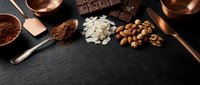 Chocolate making ingredients with cocoa powder, nuts, coconut flakes and dark chocolate bars arranged on black slate surface for confectionery preparation.