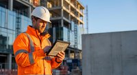 Construction worker in safety gear using tablet at building site during golden hour for project management and digital workflow documentation.