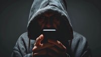 Mysterious hooded figure in darkness looking at glowing mobile phone screen, perfect for cybercrime, digital security, or online privacy concepts in modern technology articles and presentations.