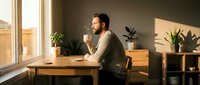 Young bearded man drinking coffee while working at wooden desk in bright modern home office with plants and natural sunlight streaming through windows.