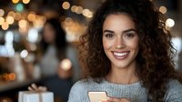 Young mixed race woman with curly hair smiling while using smartphone in cafe. Warm bokeh lights create cozy atmosphere. Casual lifestyle portrait for social media and apps.