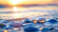 Glistening water drops on smooth pebbles at sunset create magical bokeh effect with golden sun reflection on beach, waves and ocean in background.
