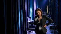 Professional businesswoman speaking into microphone at corporate event with dramatic blue stage lighting and modern venue backdrop.