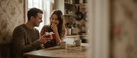 Young Caucasian couple enjoying romantic morning coffee together at kitchen table, sharing intimate conversation in cozy home interior with warm lighting.