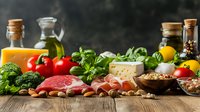 Fresh Mediterranean ingredients on rustic wooden table with raw meat, vegetables, cheese, olive oil, nuts and herbs in moody lighting setup.
