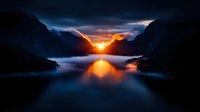 Dramatic sunset over mountain lake with golden light reflection on calm water surface between rocky peaks and low clouds creating mystical atmosphere.
