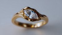 Elegant gold ring with pear-shaped diamond in unique twisted setting against neutral background, showcasing luxury jewelry craftsmanship and engagement ring design.