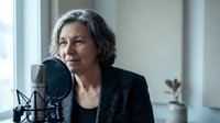 Middle-aged Hispanic woman podcaster speaking into professional microphone in bright studio setup for audio recording and broadcasting content creation.
