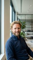 Confident middle-aged Caucasian businessman with beard smiling in modern office space with large windows and contemporary interior design for corporate use.