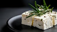 Fresh feta cheese cubes garnished with rosemary sprigs and olive oil on black ceramic plate against dark background, dramatic food photography styling.