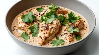 Creamy butter chicken curry with tender meat pieces in rich aromatic sauce garnished with fresh cilantro and black pepper in gray ceramic bowl on light background.