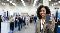 Smiling African American businesswoman holding smartphone with blank screen at busy trade show convention center, mockup ready.