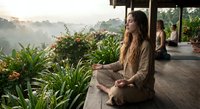 Young woman practicing mindfulness meditation on wooden deck surrounded by lush tropical plants during golden hour sunrise creating peaceful wellness atmosphere.