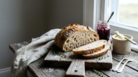 Freshly baked artisan sourdough bread sliced on rustic wooden cutting board with jam and butter, cozy kitchen window setting for food photography.