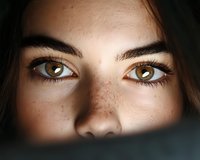 Close up portrait of young woman with freckles and expressive brown eyes in dramatic lighting, focused on eye area with dark shadows creating mysterious mood.