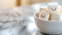 Cute marshmallows with drawn sleeping faces in white ceramic bowl on marble surface. Soft focus food photography for blogs and social media marketing.