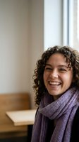 Happy young woman with curly hair wearing purple knitted scarf laughing joyfully in cozy indoor setting with natural window light for lifestyle content.