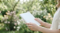 Woman holding open spiral notebook outdoors in blooming garden, wearing white shirt, writing or planning in natural sunlight.