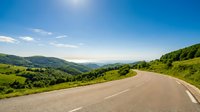 Winding asphalt road curves through lush green rolling hills and mountains under clear blue sky, suitable for travel, transportation, and landscape design projects.
