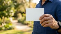 Man in blue shirt holding blank white card outdoors with green garden background, suitable for business messaging and promotional campaigns.