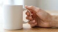 Caucasian person's hand holding white ceramic mug on wooden surface, suitable for beverage, coffee, tea, and lifestyle marketing campaigns.