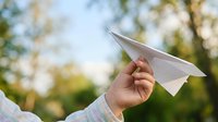 Close-up of child's hand holding paper airplane outdoors with blurred green foliage background, symbolizing freedom, imagination, and childhood play.