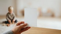 Close-up of hand holding blank white card with young child sitting in blurred background on floor, suitable for greeting cards, announcements, and family-related marketing materials.