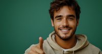 Young Hispanic man in beige hoodie showing thumbs up with confident smile against green background. Positive attitude and friendly expression for lifestyle content.