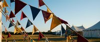 Colorful triangular bunting flags strung across outdoor festival grounds with white tents and mountains in background during golden hour sunset.