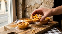 Hand placing Portuguese custard tart on rustic wooden board in traditional bakery setting with stone wall background for culinary marketing.