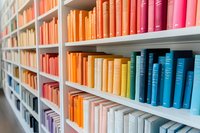 Colorful books arranged by spectrum colors on white library shelves, creating rainbow effect. Books organized in gradient from blue through orange to pink tones.