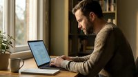 Focused businessman in brown sweater working on laptop at wooden desk by window in cozy home office with warm natural lighting and plants.