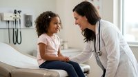 Female pediatrician examining happy African American girl patient in modern medical office during routine checkup appointment.