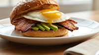 Gourmet breakfast burger with crispy bacon, fried egg, fresh lettuce on artisan bun served on white plate for restaurant menu and food advertising.