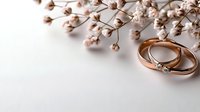 Elegant rose gold wedding rings with diamond accent beside delicate dried baby's breath flowers on white background for romantic ceremony planning.