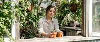 Woman in casual beige shirt arranging small potted plants on wooden table in bright greenhouse surrounded by green foliage and natural light.