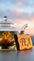 Glass teapot with blooming flower tea and rustic wooden TEA sign against soft pastel sky, cozy herbal drink concept.