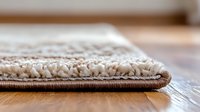 Close up view of carpet edge showing plush cream colored pile texture and brown binding on wooden floor, demonstrating quality flooring materials and installation.