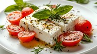 Fresh feta cheese block with sliced tomatoes, rosemary sprigs and black pepper on white plate for Mediterranean cuisine, salad recipes and healthy food styling.