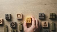 Hand placing a glowing wooden block with target icon among business symbol cubes on wooden surface, representing strategy and goal achievement.