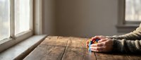 Person holding warm coffee mug at rustic wooden table by window with natural light creating cozy morning atmosphere for relaxation and comfort.