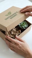 Female hands opening kraft paper box with green leaf logo containing succulent plant wrapped in tissue paper, showcasing eco-friendly packaging for sustainable product delivery and unboxing experience.
