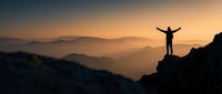 Silhouette of person with raised arms celebrating success on mountain peak at sunrise with layered hills and golden sky for motivation concepts.