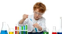 Young redhead male scientist in lab coat and safety goggles conducting chemical experiment with colorful test tubes and beakers on white background.