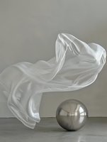 Flowing white silk fabric floating above metallic sphere on gray background creates dynamic contrast between soft textile movement and solid geometric form.