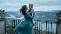 Mother in teal dress holding toddler son on mountain terrace overlooking valley landscape. Family bonding moment with scenic backdrop for lifestyle content.