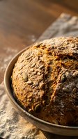 Freshly baked artisan bread loaf with crusty golden-brown exterior resting in ceramic bowl on flour-dusted wooden surface, ideal for bakery, food, and culinary content.