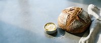 Freshly baked artisan bread loaf with crusty exterior next to butter in small bowl on blue surface with white cloth, ideal for bakery, food, and culinary content.